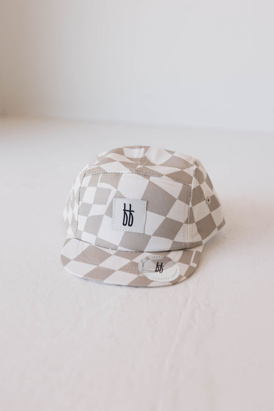 A beige and white checkered Children's Snap | Wavy Checker hat by forever french baby, featuring a white rectangular patch with black lowercase letters "bb," is centered on a white surface. Designed for ages 4 months to 6 years, this accessory also includes a smaller similar patch sewn on the front of the brim.