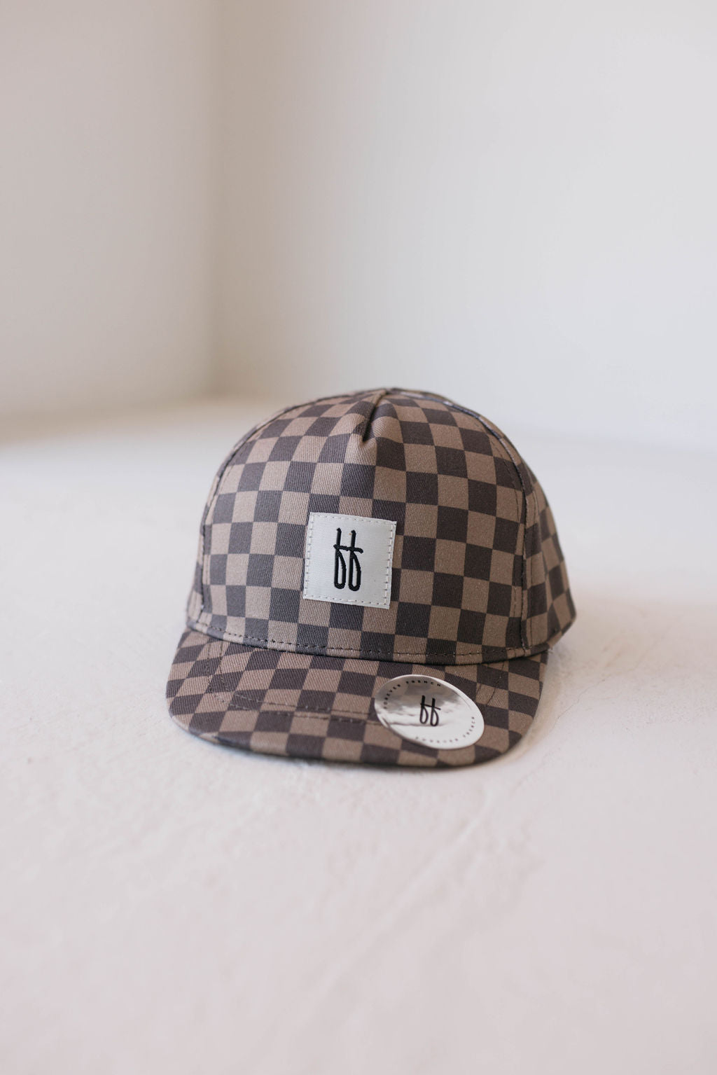 A Children's Trucker Hat by forever french baby is displayed on a white surface. Featuring a faded brown checkerboard pattern, it has a small rectangular patch with the letters "bb" in black centered on the front and offers good sun protection. A round sticker adorns the brim.