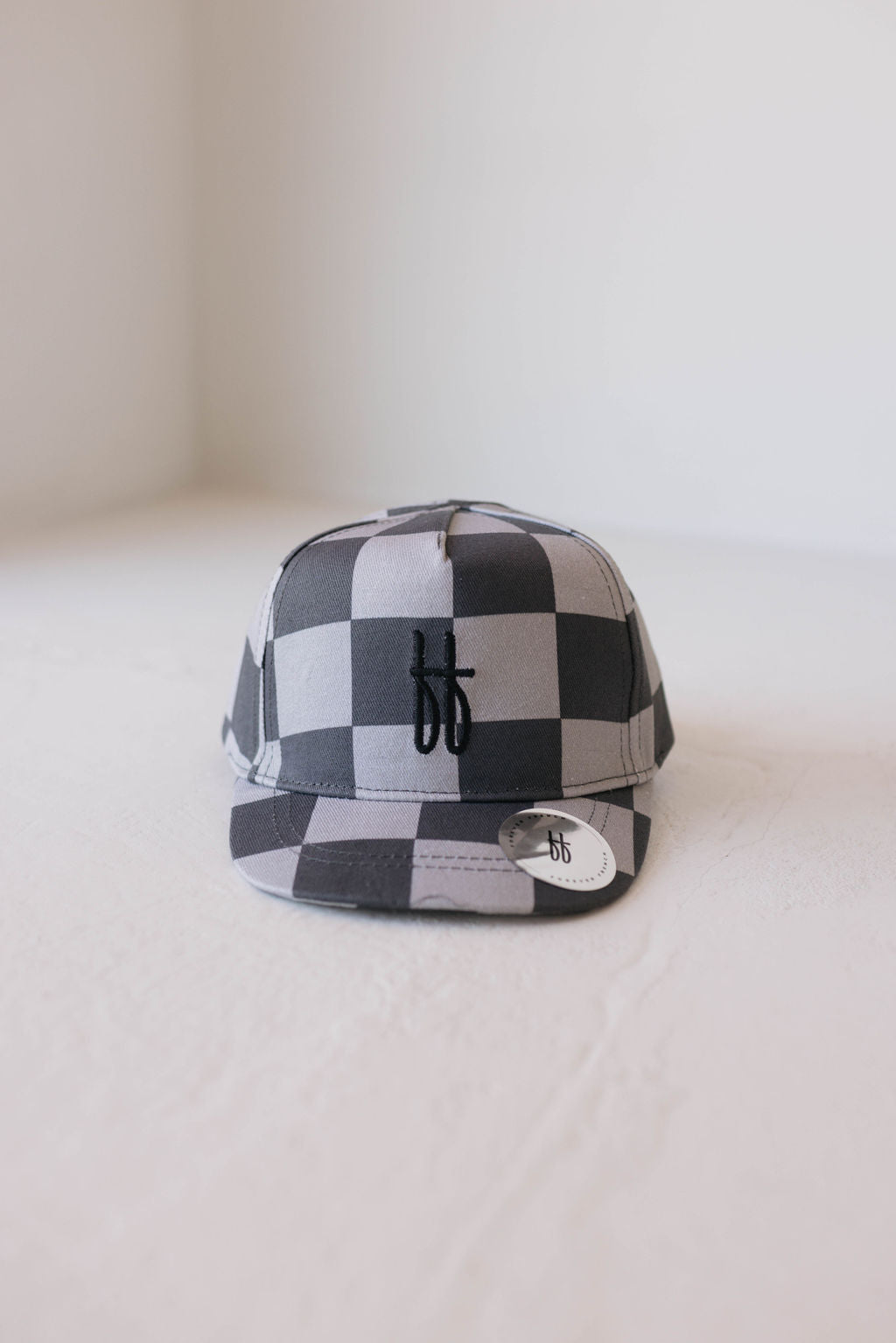 A Children's Trucker Hat with a black and gray checkerboard pattern from forever french baby, featuring a small "bb" logo on the front and a round sticker on the brim, ideal for sun protection, sits on a white surface against a plain, light-colored wall.