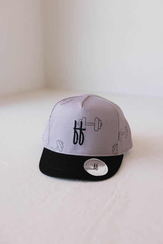 A Children's Trucker Hat in gray with a black brim from forever french baby is depicted. This "Arm Day" hat showcases a pattern of outlined dumbbell illustrations and an embroidered "ff" logo on the front panel. Designed for sun protection, it also features a sticker with the same "ff" logo on the brim. The background is a plain, light-colored surface.