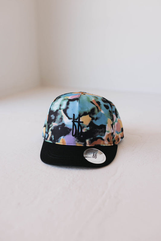 The Children's Trucker Hat from forever french baby showcases a vibrant Charli Print tie-dye design, featuring shades of blue, green, yellow, and pink. It is complemented by a black bill and adorned with a small black logo on the front. This inspired cap, which draws influence from autistic child art, is displayed resting on a light-colored surface with an additional sticker displaying the same logo on the bill.