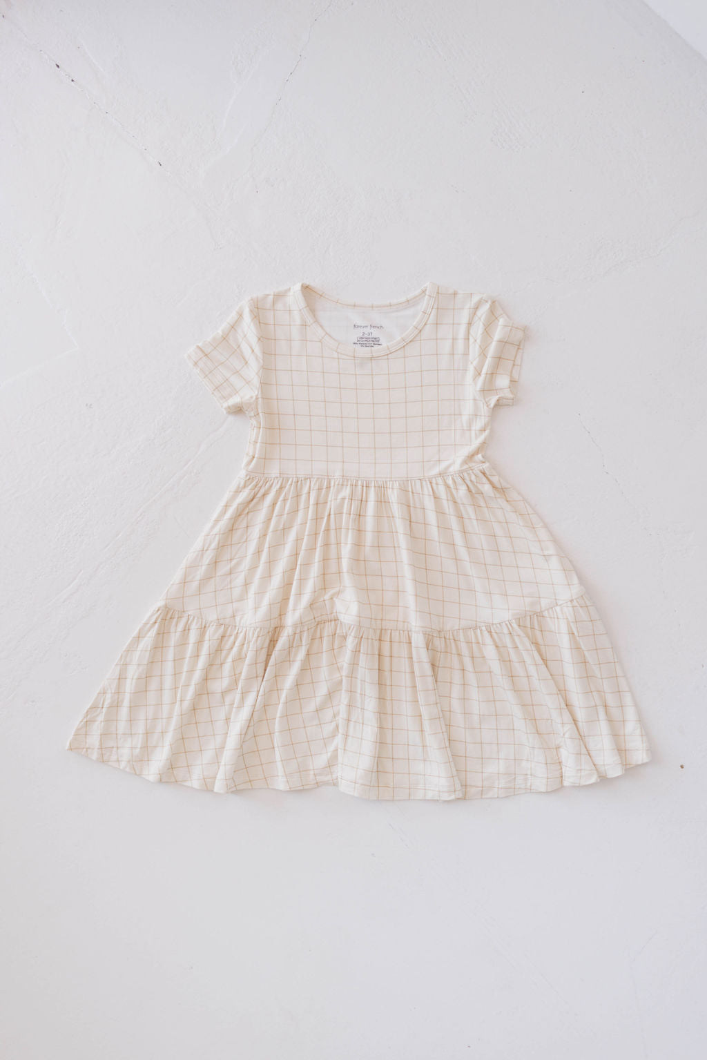 A Multi Tier Bamboo Dress | Golden Grid from forever french baby, featuring a cream color and short sleeves, is laid out on a white surface. It has a round neckline and a tiered skirt that gives it a ruffled appearance, combining stylish design with sustainable bamboo fabric.