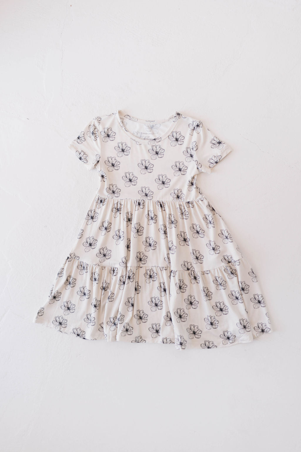 The Multi Tier Bamboo Dress by forever french baby in Desert Bloom features a cream-colored, short-sleeve, knee-length design with a gathered waist and a flared skirt. It is adorned with black floral outlines and made from sustainable bamboo fabric, combining eco-friendly charm with a light-colored surface.