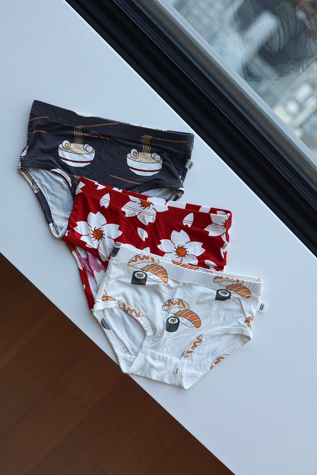 Three pairs of forever french baby Girl's Bamboo Briefs (3 Pack)—featuring Cherry Blossoms, Ramen, and Sushi prints—are displayed on a white surface; hypoallergenic girls’ underwear offering comfort and playful style.