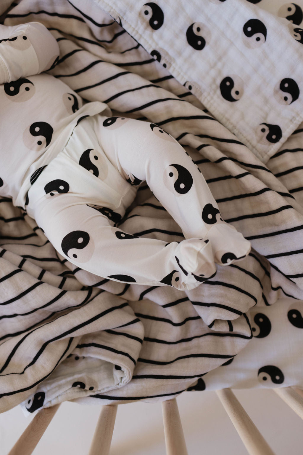 A baby is lying on a bed covered with black and white striped blankets, wearing hypo-allergenic clothing adorned with a black Yin Yang symbol pattern. The baby is partially covered by another blanket featuring the same Yin Yang design from the Sleepy Time Set by Forever French Baby.