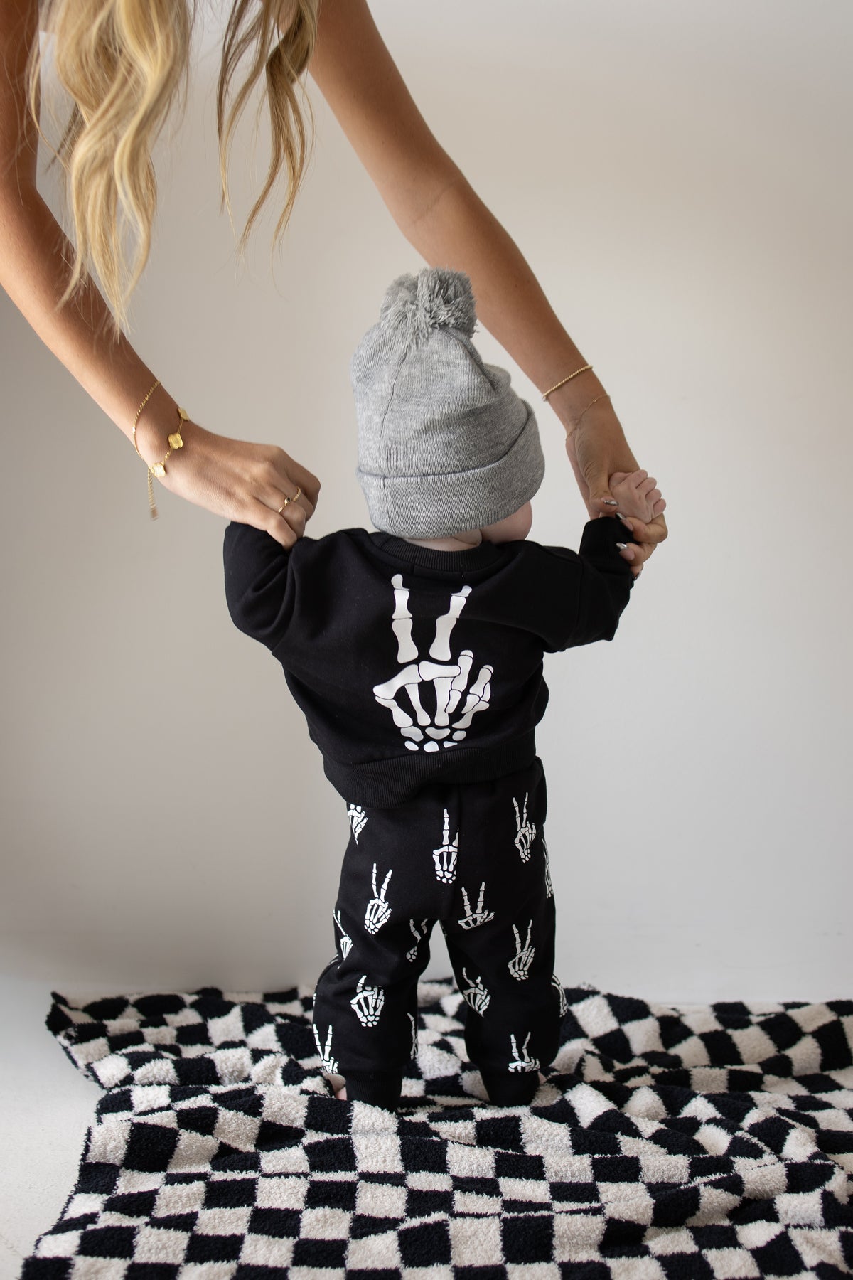 A baby wearing the forever french baby Child Sweat Set | Midnight Skelly and a gray beanie stands on a black-and-white rug, holding hands with an adult whose face is out of frame—ready for everyday adventures.