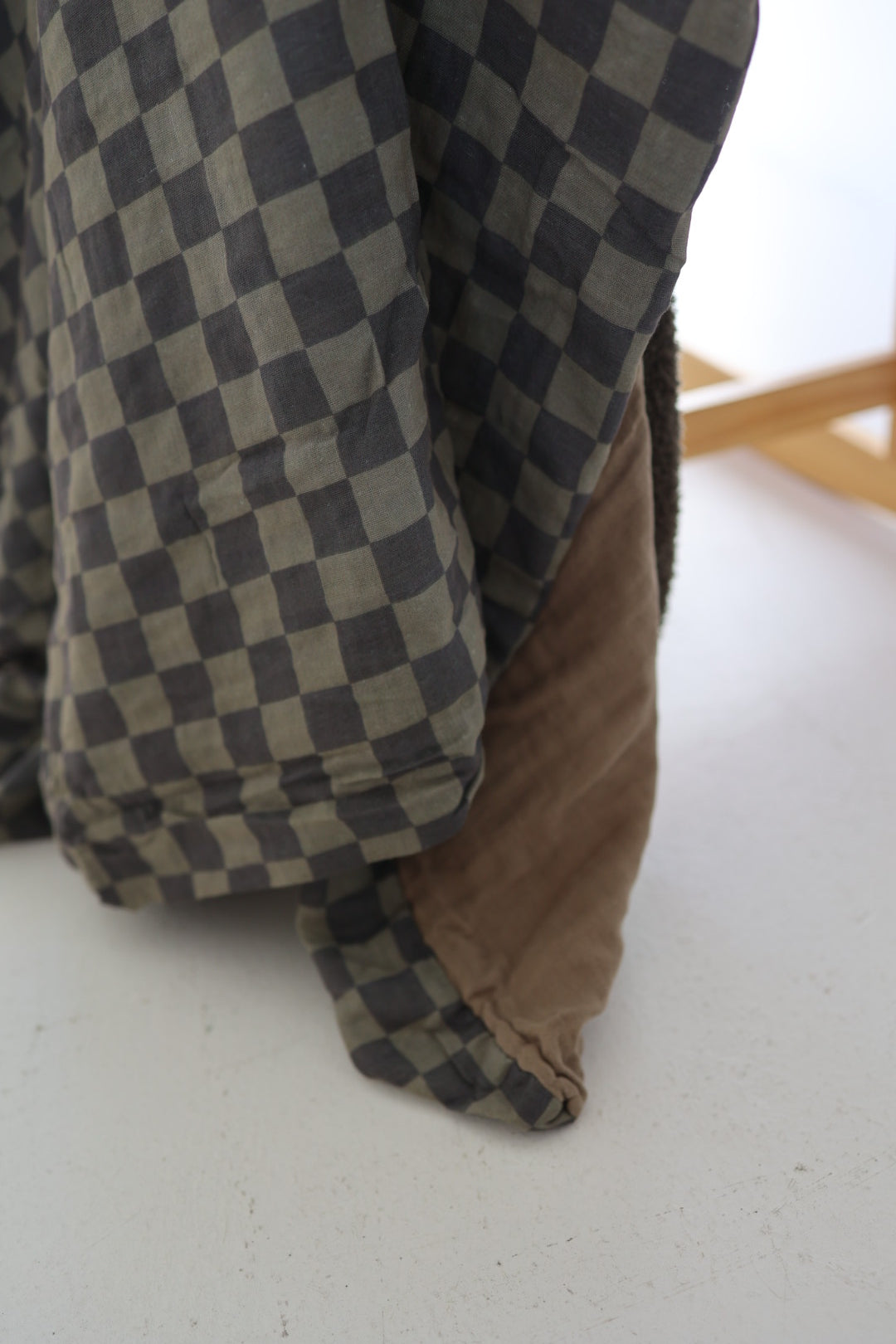The Muslin Quilt | Olive Checkerboard by forever french baby features dark and light gray squares, draped over soft brown fabric—ideal for cozy nursery bedding on a light background.