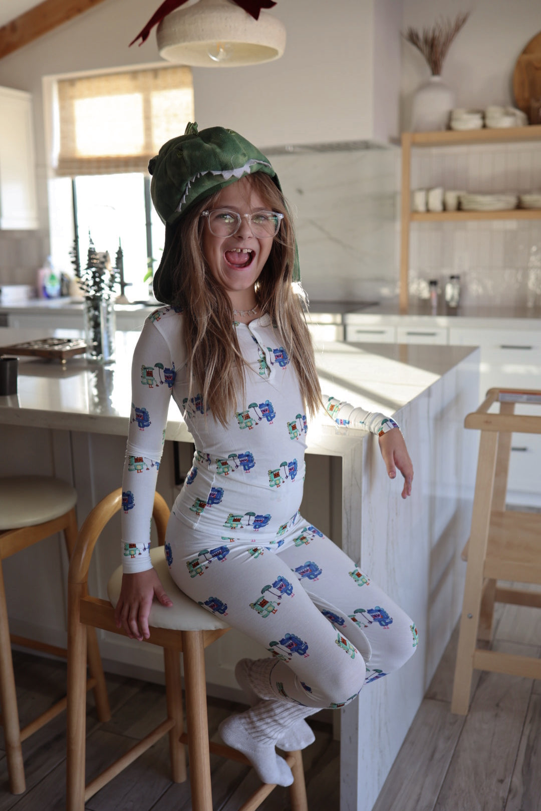 A cheerful girl in glasses and a green dinosaur hat sits on a kitchen stool, wearing forever french baby’s Bamboo Two Piece Pajamas | Charli's Monsters, with sunlight streaming into the modern kitchen behind her.