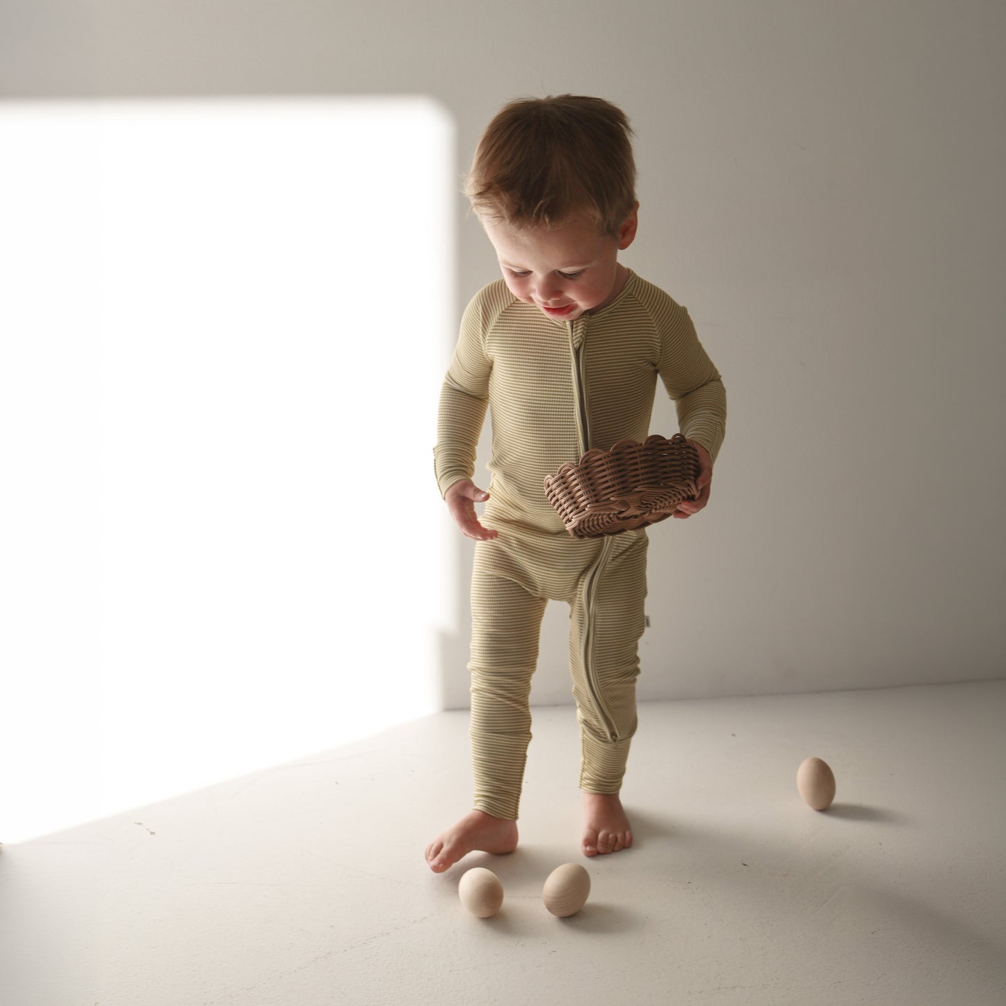 A small child in forever french baby Bamboo Zip Pajamas | Willow Stripe Ribbed stands barefoot on a white floor, holding a woven basket and looking down at three wooden eggs in soft natural light.