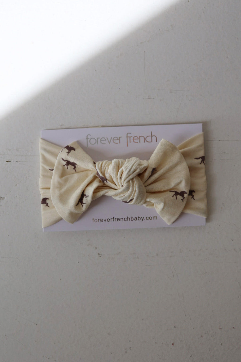 The Bamboo Head Wrap | Wild Horses by forever french baby is a cream bamboo baby headband with a center knot and brown horse prints, displayed on a "forever french baby" card with "foreverfrenchbaby.com" visible.