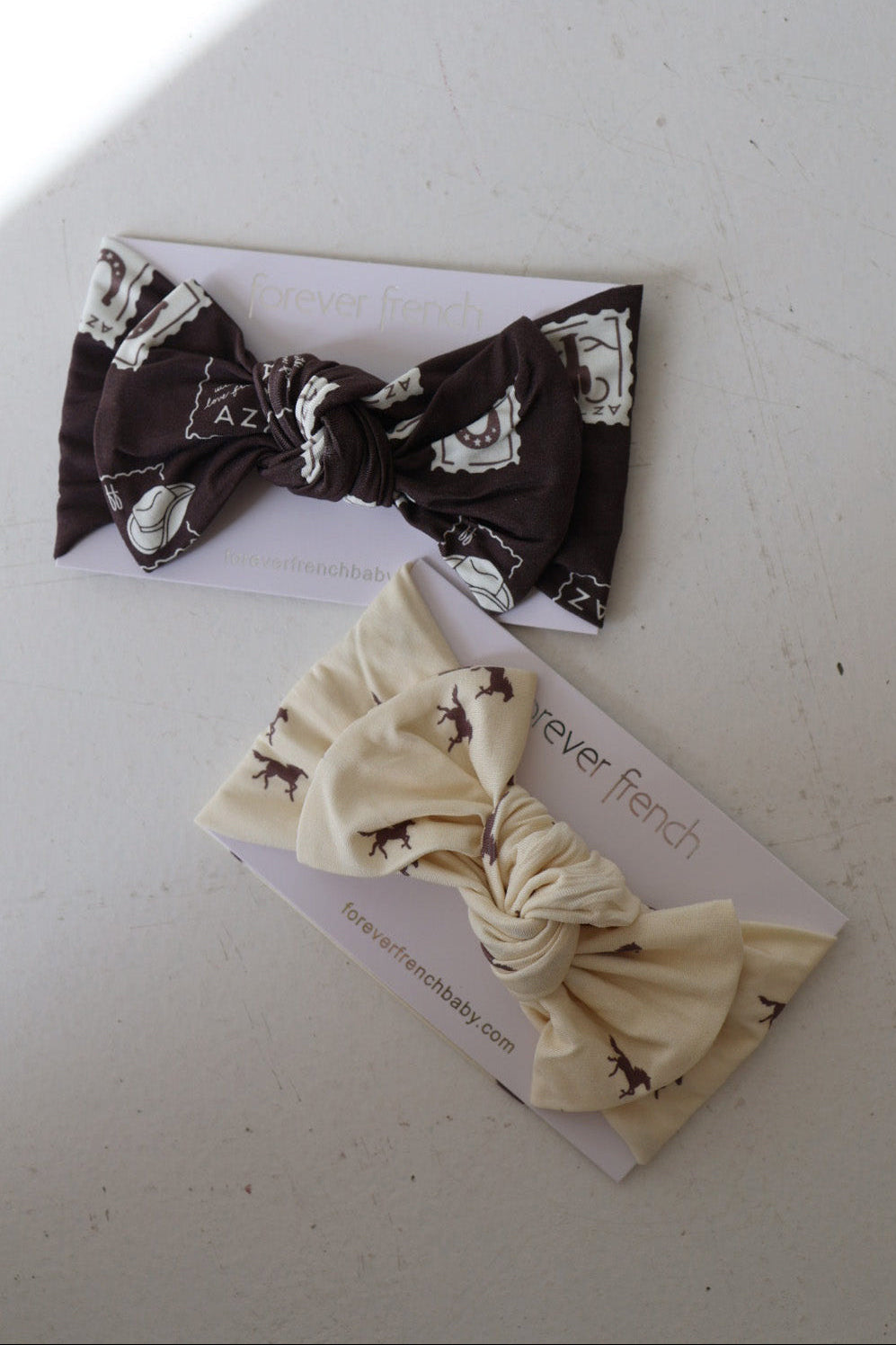 The Bamboo Head Wrap by forever french baby includes two knotted headbands: a dark band with white patterns and a cream band with brown animal prints. Displayed on branded cards, they add a stylish touch for your little one.