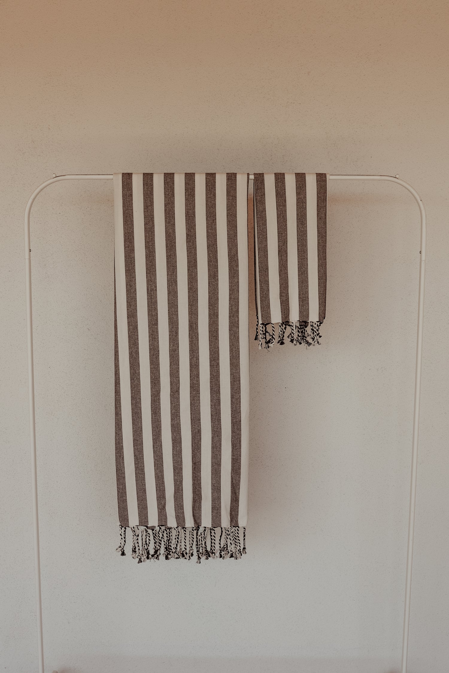 A white towel rack holds two forever french home Black Stripe Turkish Towels, one large and one small. Each towel features vertical brown and black stripes adorned with fringe tassels at the ends. The plain, light-colored wall in the background perfectly highlights the elegance of these meticulously crafted towels.