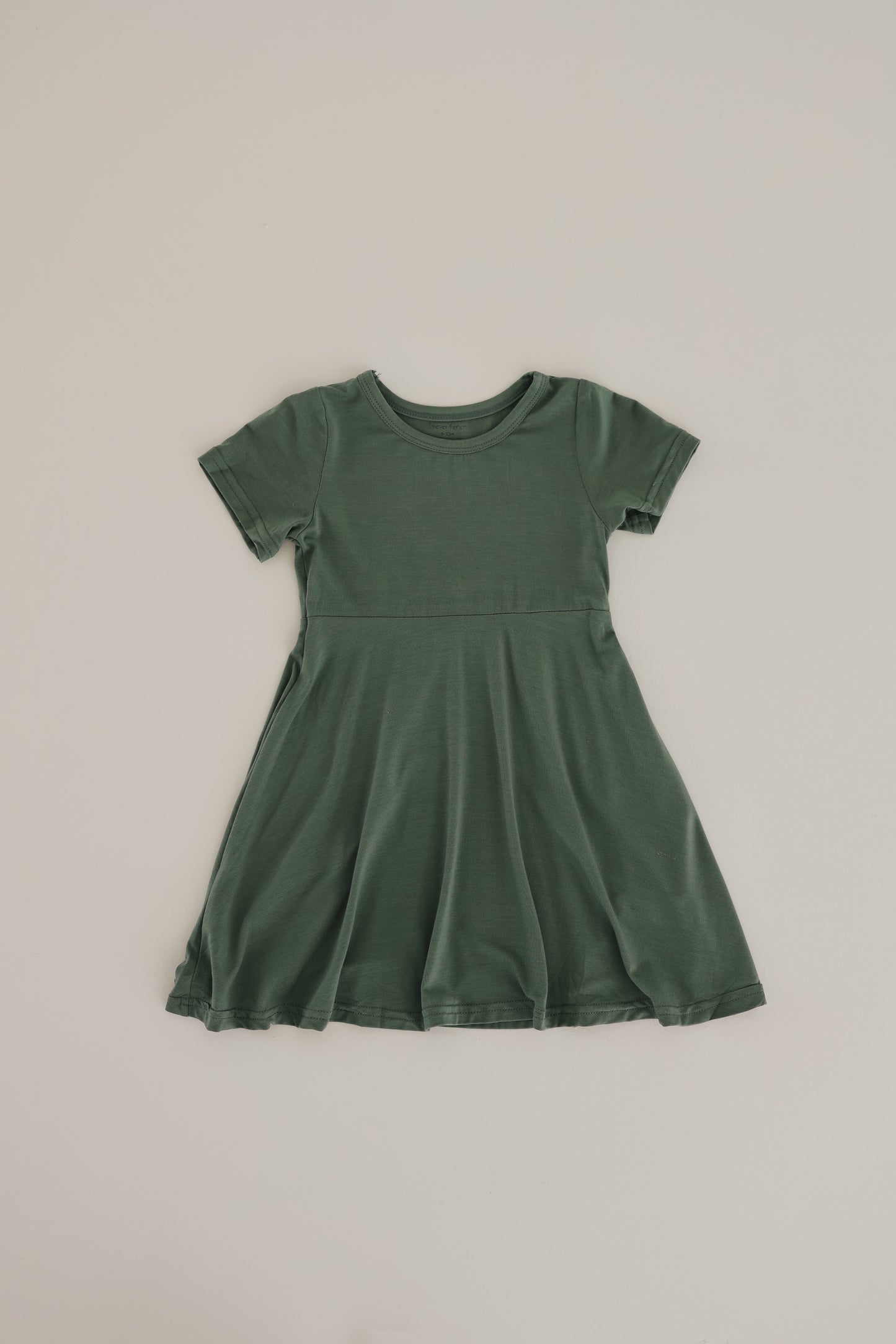 A child-sized, short-sleeved Bamboo Dress in Moss from the forever french baby collection is laid flat against a light beige background. The dress boasts a fitted bodice and a flared, twirl-worthy skirt, offering a simple yet charming design. Perfect for those seeking hypo-allergenic clothing.