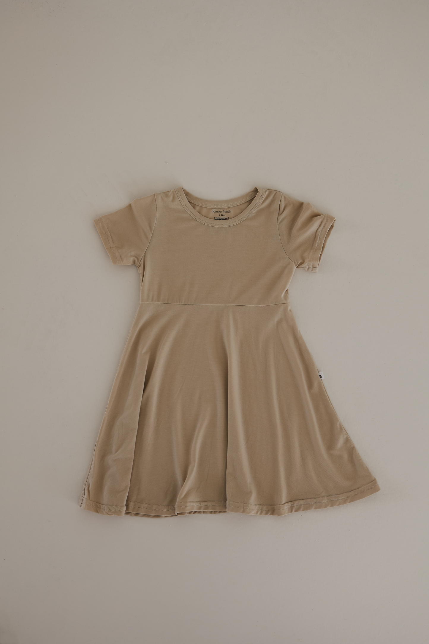 The Bamboo Dress in Oatmeal by forever french baby is a short-sleeved, knee-length design laid flat against a plain background. It's breathable with a round neckline and slightly flared skirt, making it perfect for hypo-allergenic clothing fans.