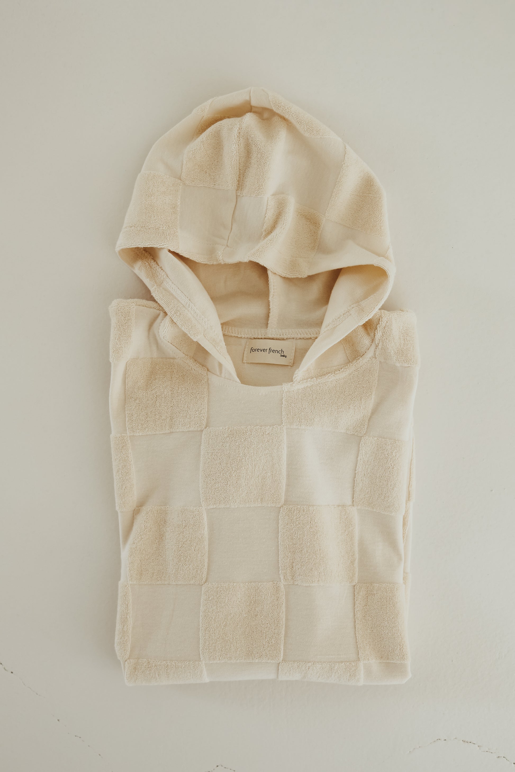 A Hooded Towel in a cream checkerboard pattern is laid flat on a light surface. The towel showcases a combination of fleece and smooth textures in a patchwork design. A small tag near the corner displays the forever french baby brand label.