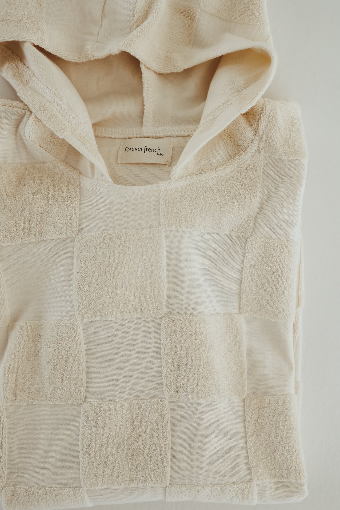 A folded cream-colored hooded towel with a checkerboard design and a label inside the collar reading "forever french baby." The fabric features a mix of smooth and textured squares, creating a unique look. The towel is laid on a light surface, resembling luxurious bath towels in its softness.