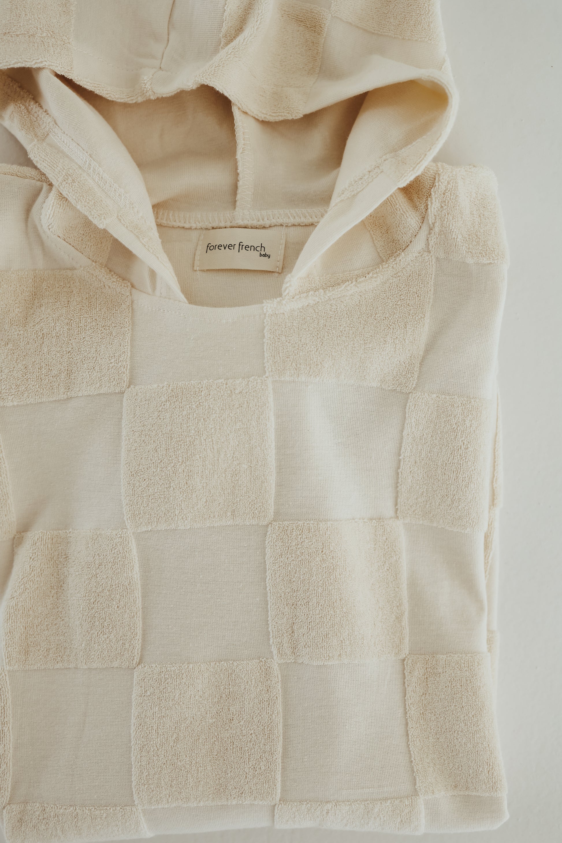 A folded cream-colored hooded towel with a checkerboard design and a label inside the collar reading "forever french baby." The fabric features a mix of smooth and textured squares, creating a unique look. The towel is laid on a light surface, resembling luxurious bath towels in its softness.