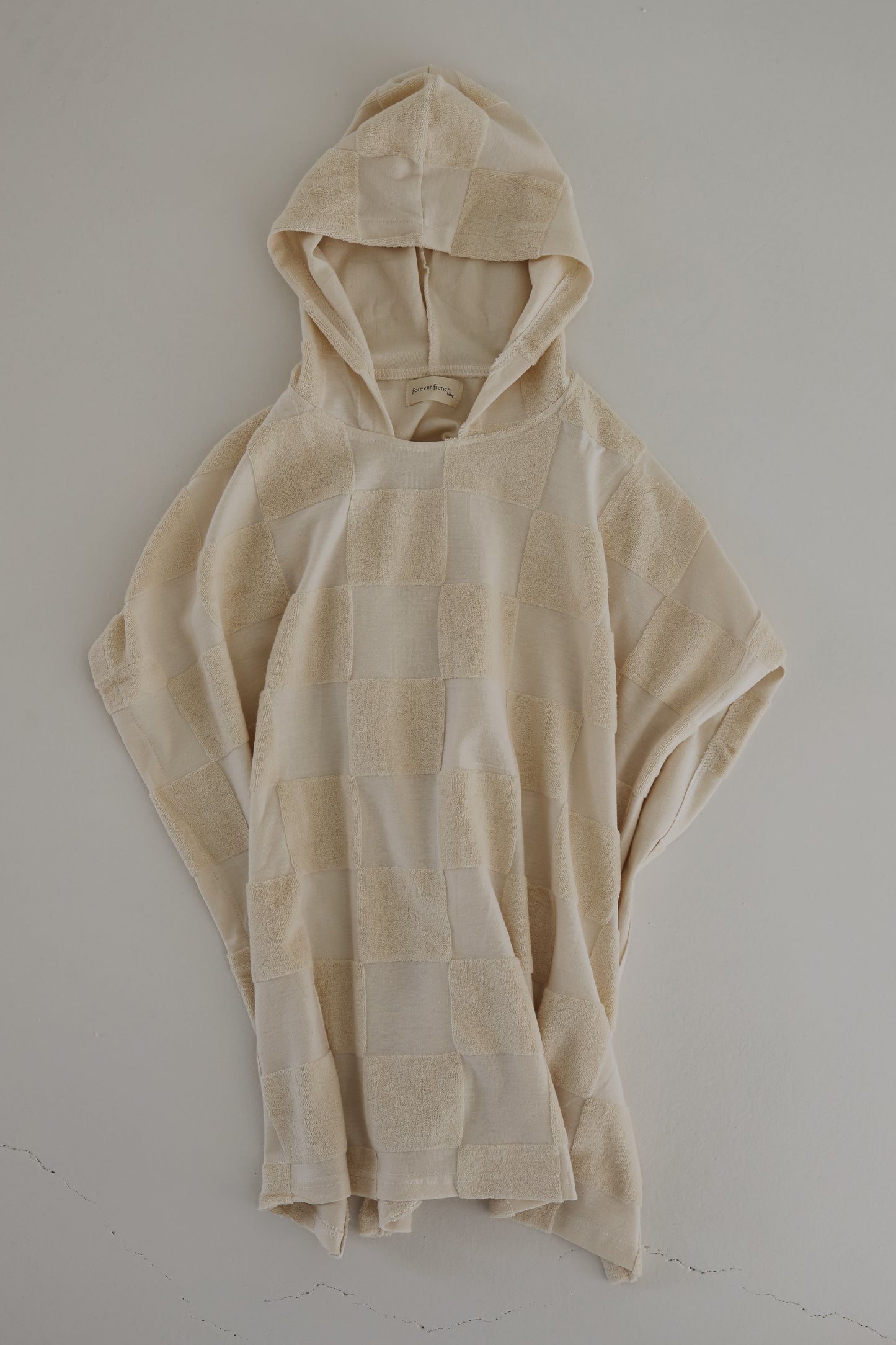 The Hooded Towel in Cream Checkerboard by forever french baby features a soft and textured fabric with a checkerboard design in varying shades of beige. Designed for comfort, it has a loose and relaxed fit with a casually draped hood that gives the towel a cozy and laid-back appearance.