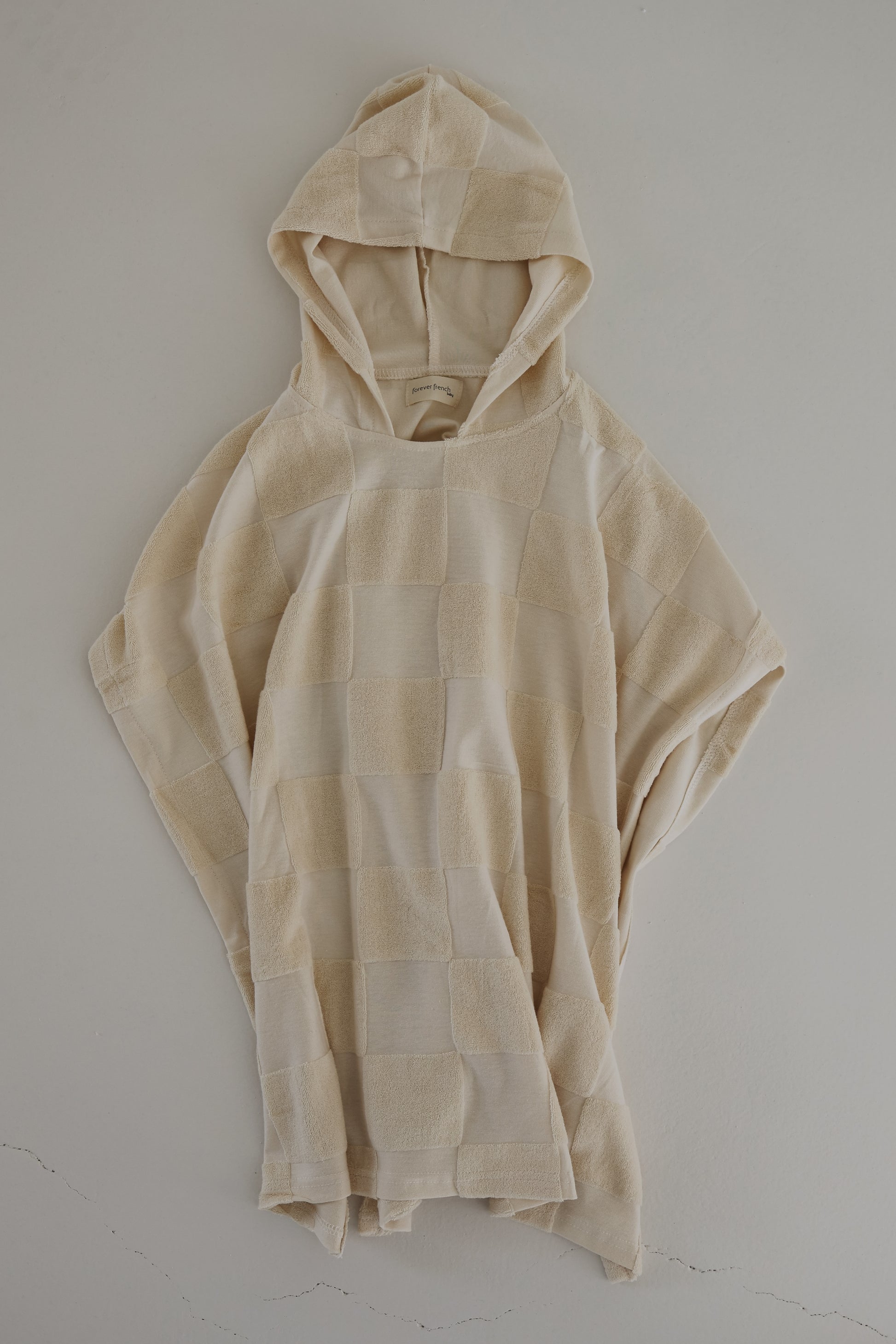 The Hooded Towel in Cream Checkerboard by forever french baby features a soft and textured fabric with a checkerboard design in varying shades of beige. Designed for comfort, it has a loose and relaxed fit with a casually draped hood that gives the towel a cozy and laid-back appearance.