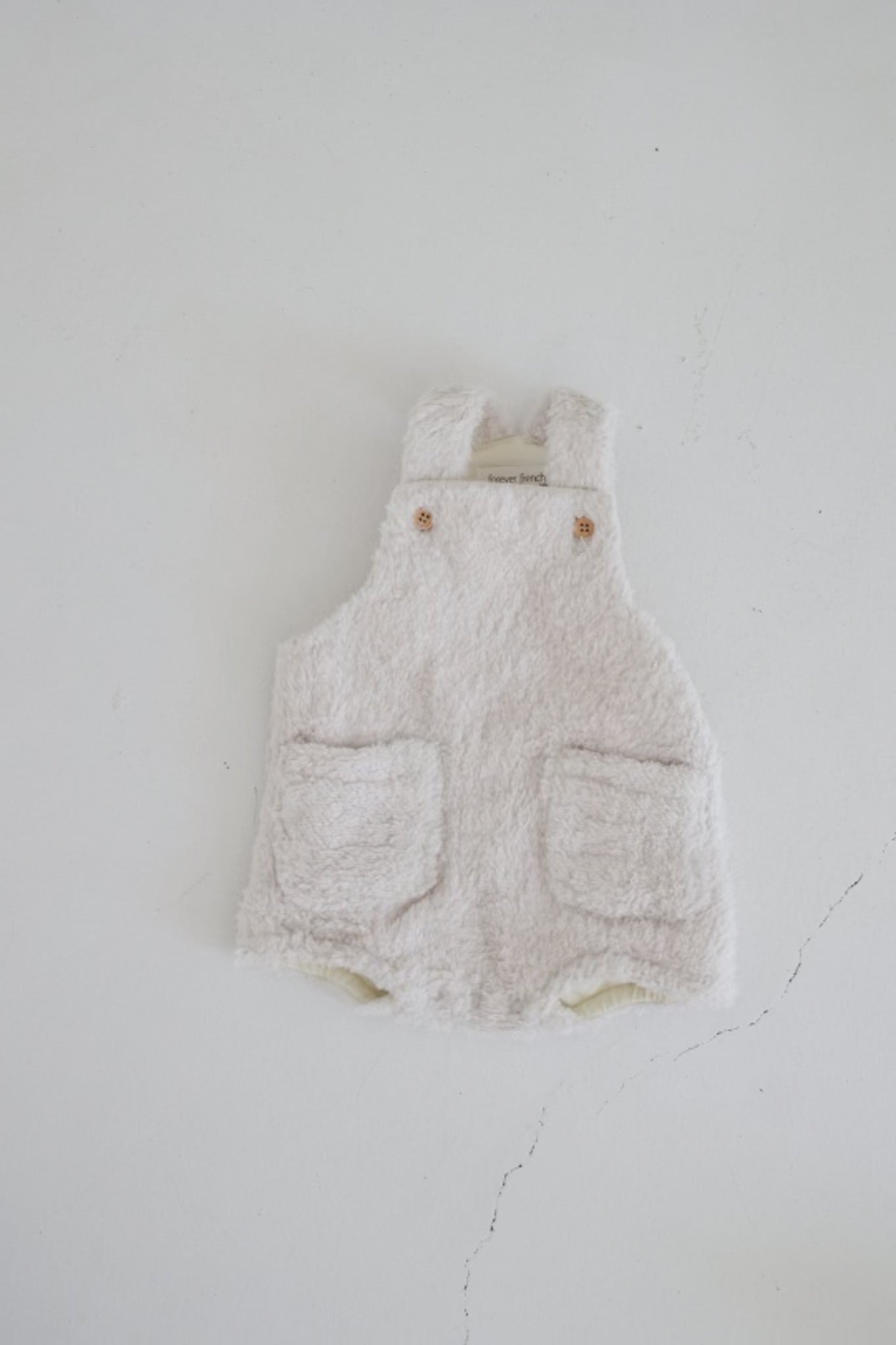 The Teddy Overalls in Sand from Forever French Baby is a soft, fluffy white baby romper that features two front pockets and button closures at the shoulders. Laid flat on a light surface, this piece has a cozy, textured appearance reminiscent of classic teddy overalls in a timeless neutral sand color.