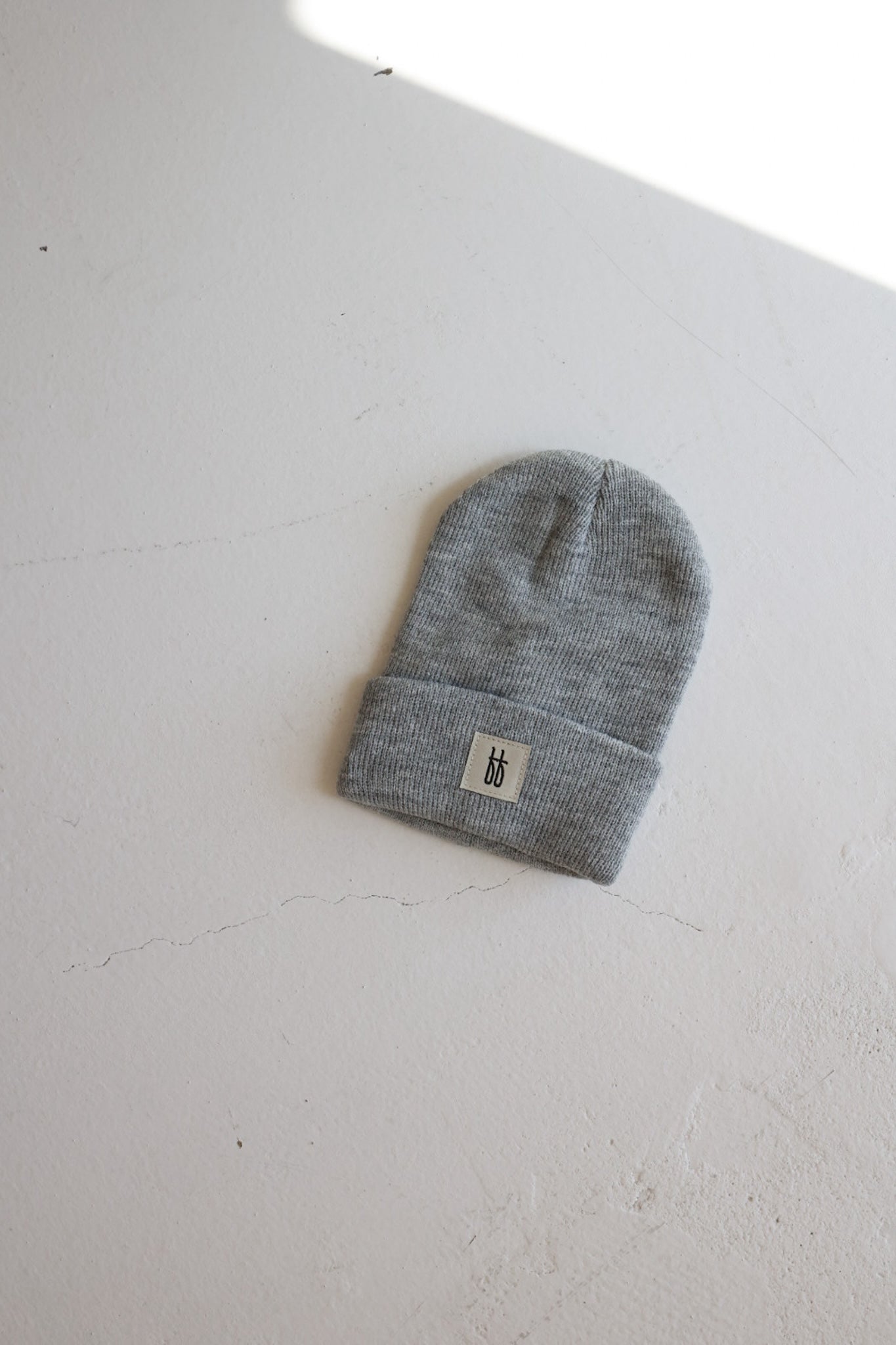 The forever french baby ff Knit Beanie in gray lies flat on a light-colored surface. This classic ribbed design features a simple, folded brim with a small rectangular label displaying two vertical black lines in the center.