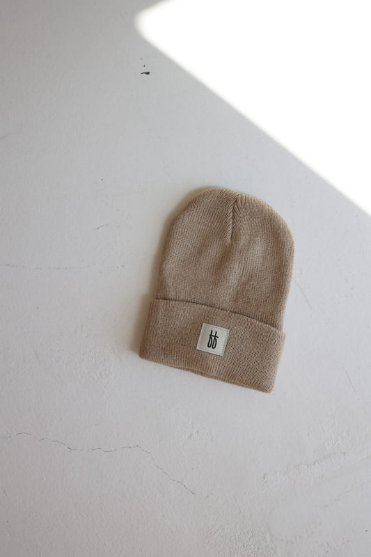 A taupe knit beanie from the Forever French Baby collection, featuring a folded brim and a small rectangular white patch with the letters "ff" in black, is placed on a light-colored surface with a shadow cast across part of it.