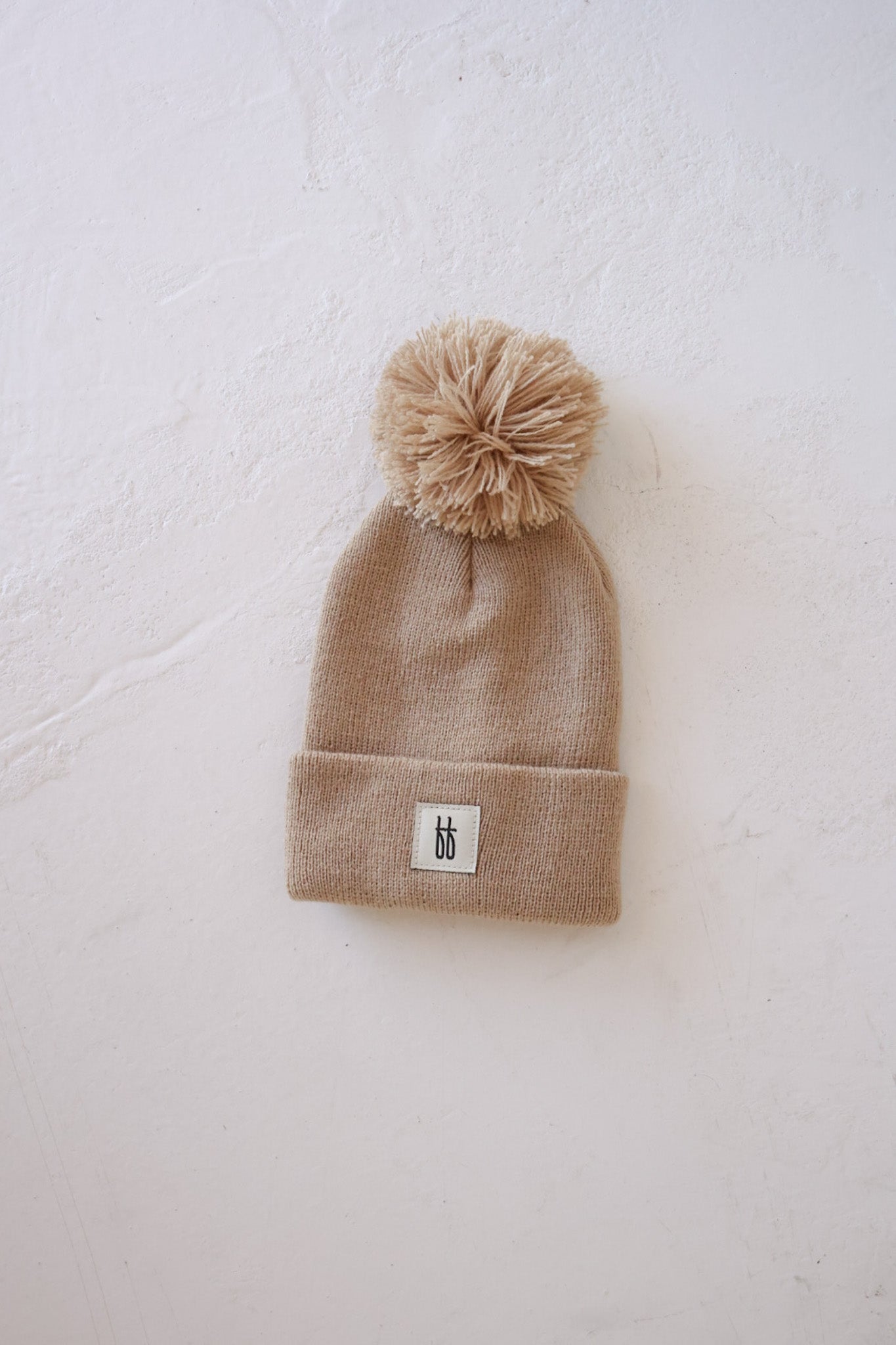 A taupe ff Knit Pom Top Beanie by forever french baby, featuring a fluffy pom-pom on top and a folded brim with a small white rectangular patch displaying a black design on the front. The classic ribbed design brings timeless style to this cozy accessory, showcased against a light textured surface.