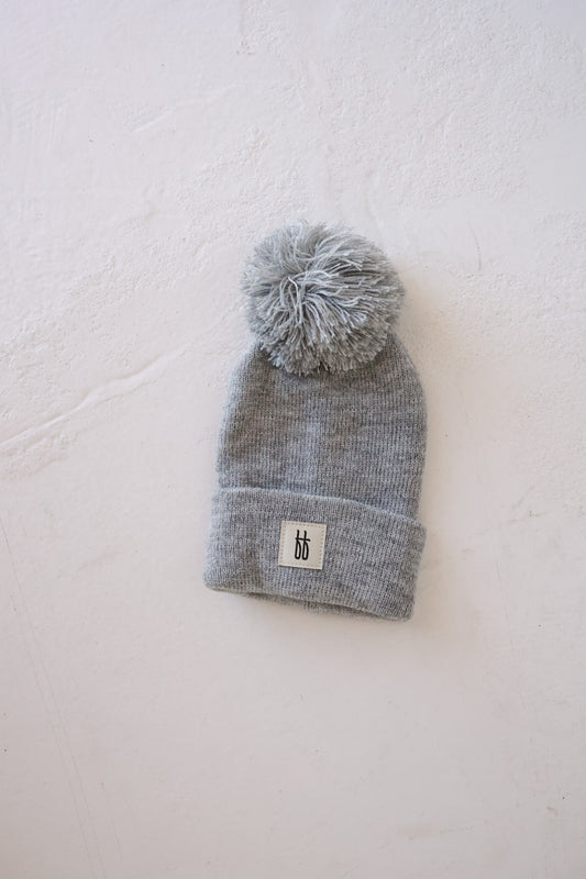 The ff Knit Pom Top Beanie in light gray from forever french baby, with a large pom-pom on top and a ribbed folded brim adorned with a small white patch featuring the letters "bb," rests on a white textured surface. This charming accessory is ideal for adding a touch of Forever French Baby style to your little one's outfit.