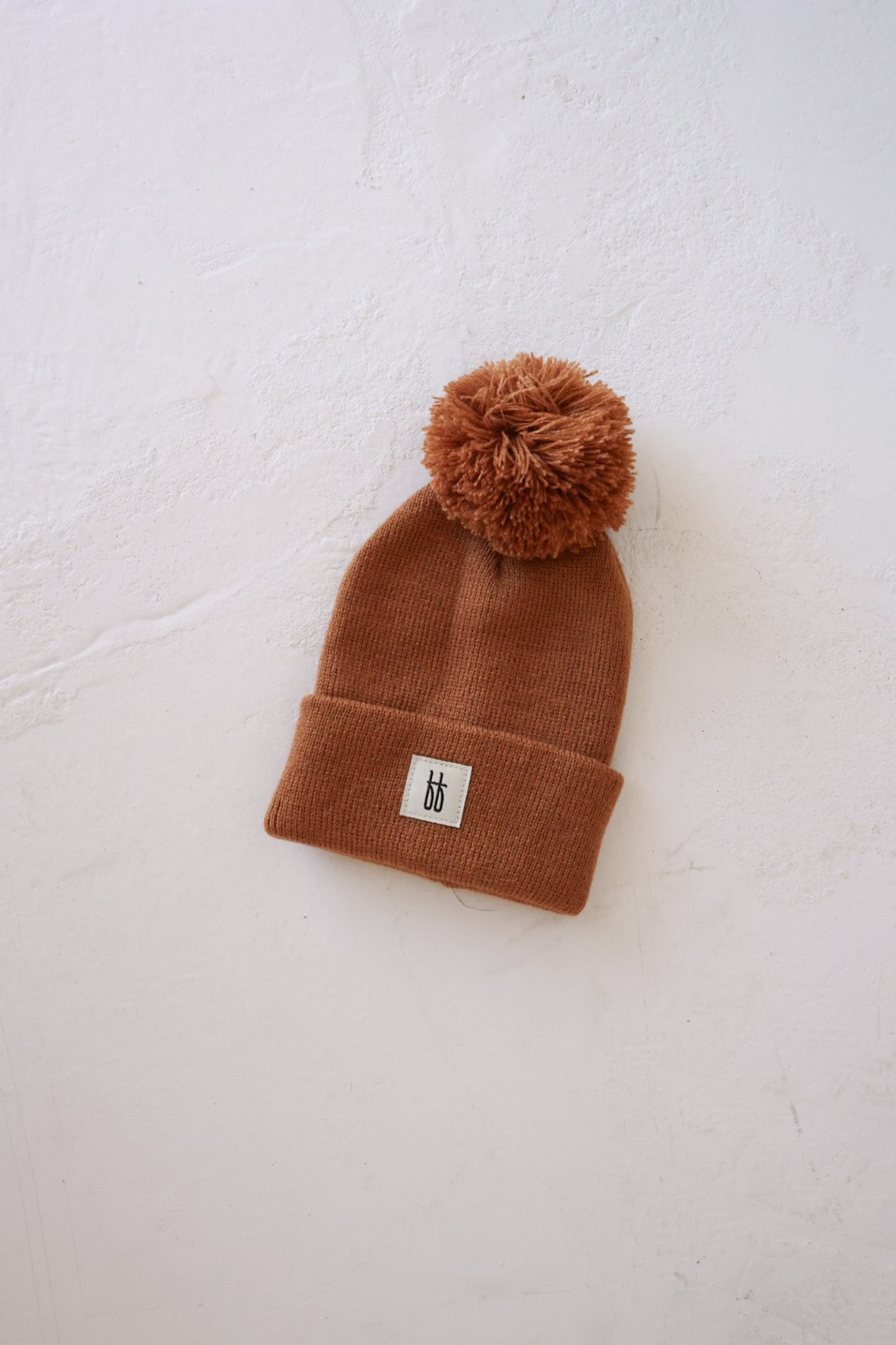 The ff Knit Pom Top Beanie in rust by forever french baby is showcased against a white textured background. This brown beanie hat features a large pom-pom on top, and the signature design includes a folded cuff adorned with a small white rectangular patch with black lettering, all crafted from breathable yarn for comfort and style.