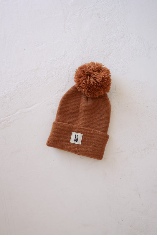 The ff Knit Pom Top Beanie in rust by forever french baby is showcased against a white textured background. This brown beanie hat features a large pom-pom on top, and the signature design includes a folded cuff adorned with a small white rectangular patch with black lettering, all crafted from breathable yarn for comfort and style.