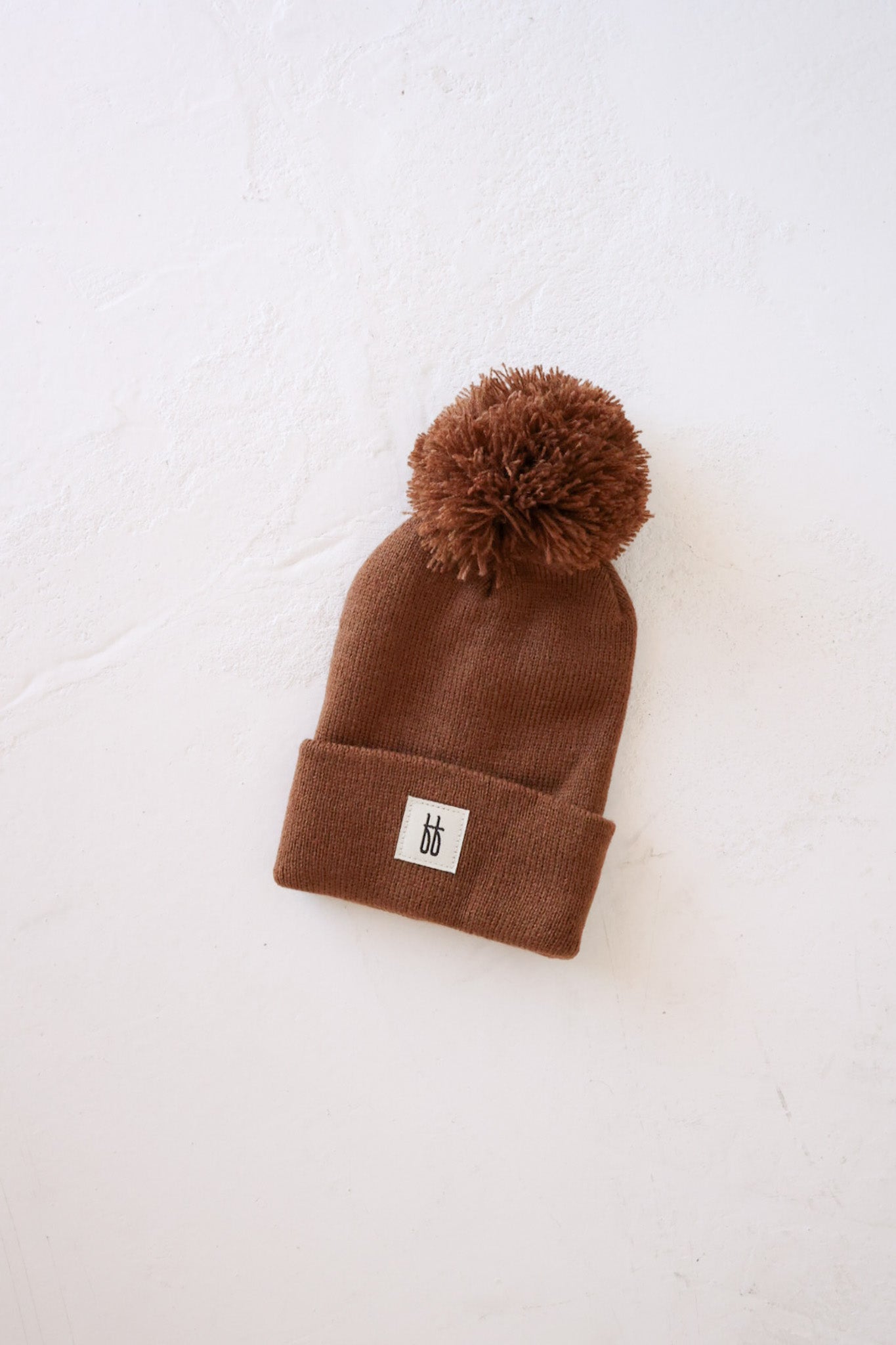 The ff Knit Pom Top Beanie in chestnut from forever french baby is laid flat on a light-colored surface. This beanie features a large pom-pom on top and a small rectangular cream-colored patch with two lowercase "b" letters in black, stitched at the folded brim.