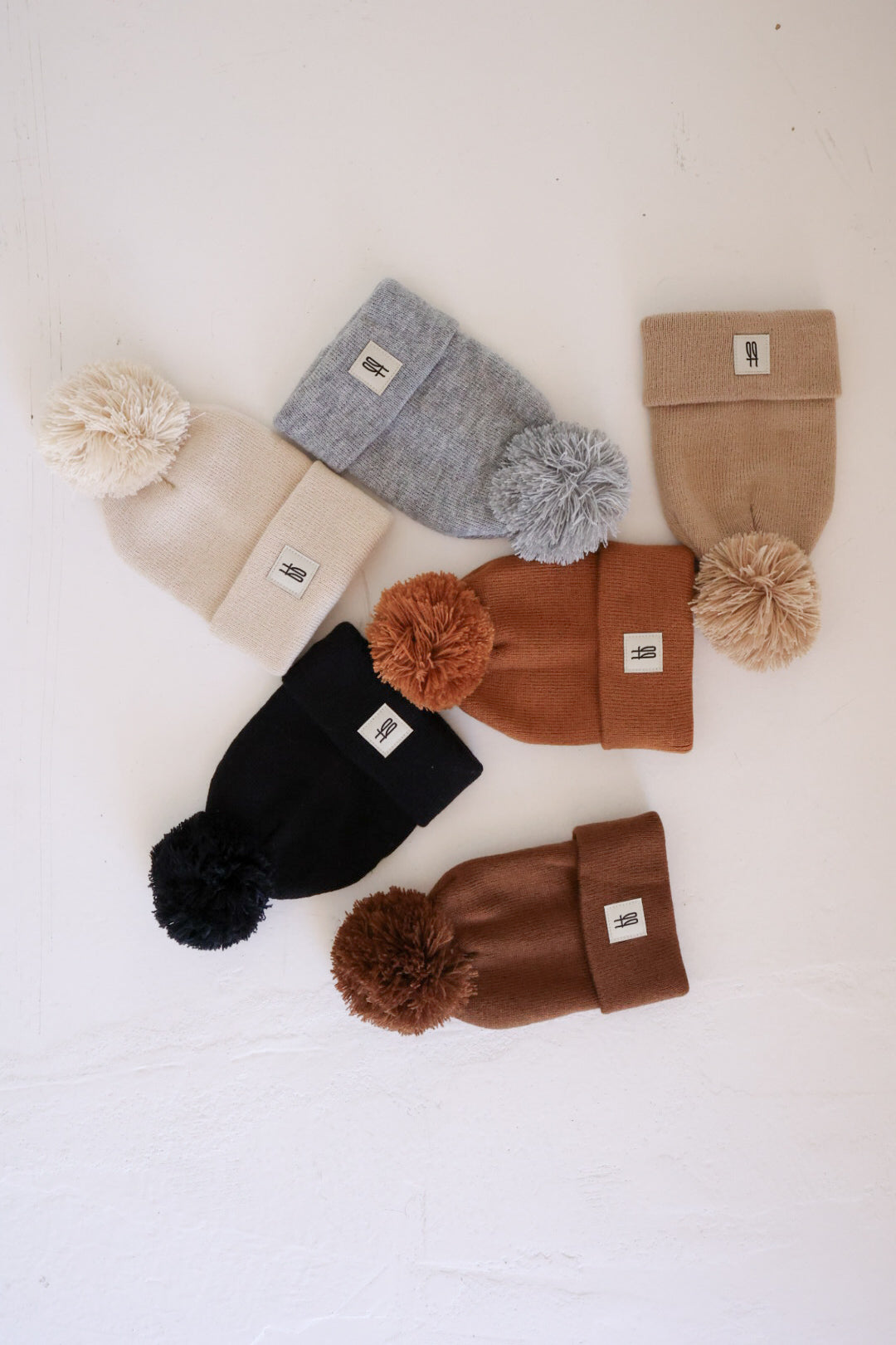 Six classic, ribbed design ff Knit Pom Top Beanies by Forever French Baby are displayed in a circular arrangement on a white surface. Each beanie features a large pom-pom on top and a small rectangular patch on the front. The collection includes an array of timeless colors such as beige, light gray, tan, black, rust, and burnt orange.