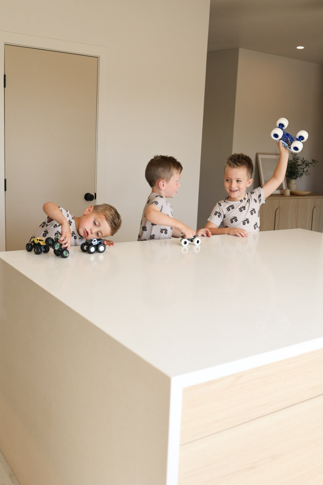 Three young boys in matching forever french baby Bamboo Short Set | Monster Truck pajamas play with toy cars on a bright white kitchen island, two standing and one leaning in, all focused on their toys in a modern room.