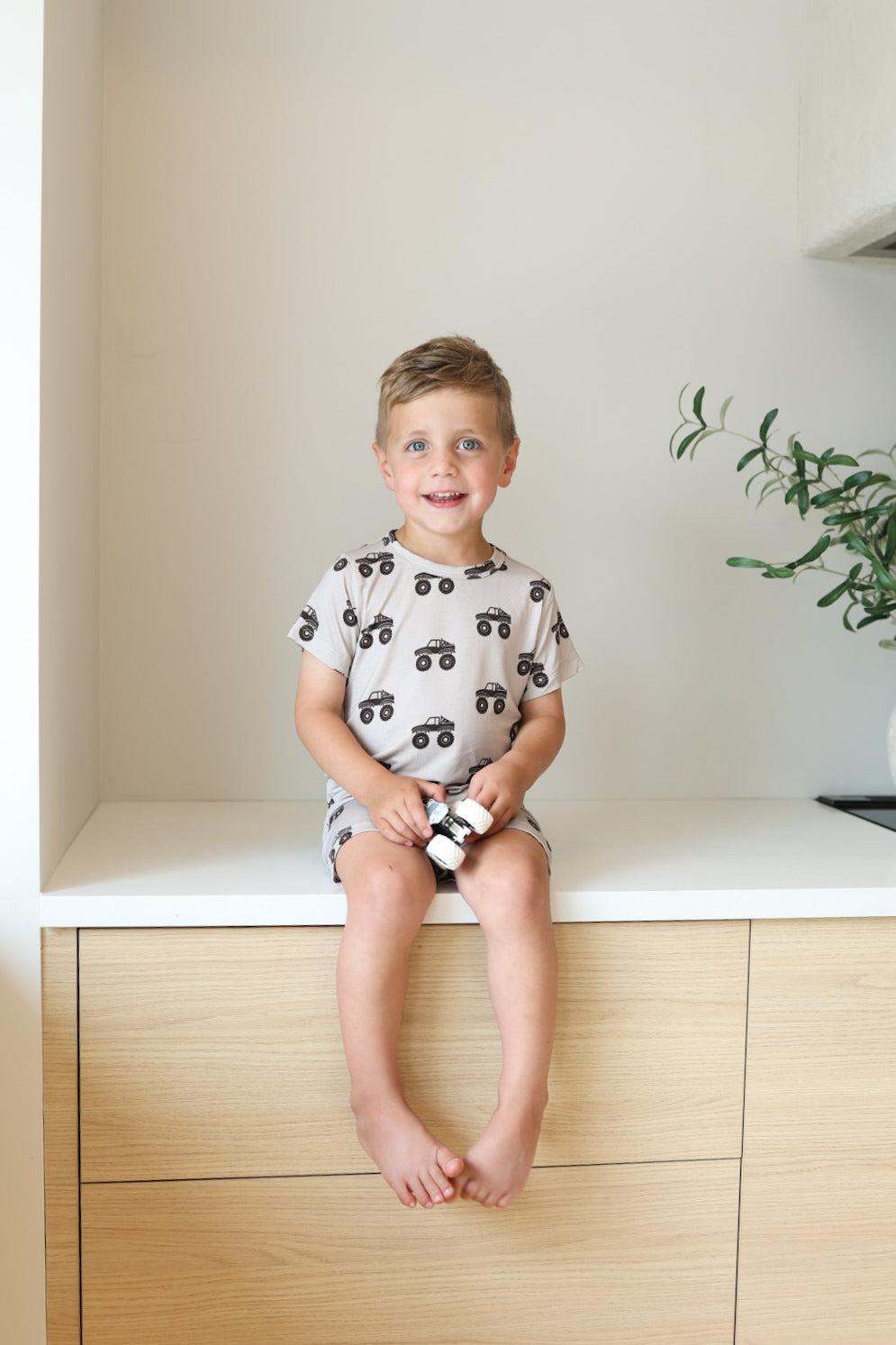 A young boy sits barefoot on a light wooden cabinet, smiling. He wears the forever french baby Bamboo Short Set | Monster Truck and holds a small toy. A green plant is visible to his right.