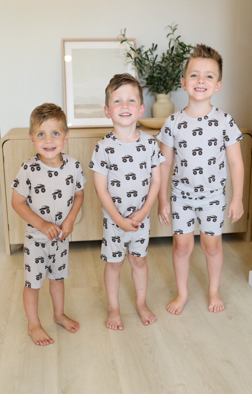 Three young boys smile at the camera indoors, wearing forever french baby Bamboo Short Set | Monster Truck—a breathable grey pajama set with black vehicle prints. A light wood cabinet, framed art, and a plant are in the background.