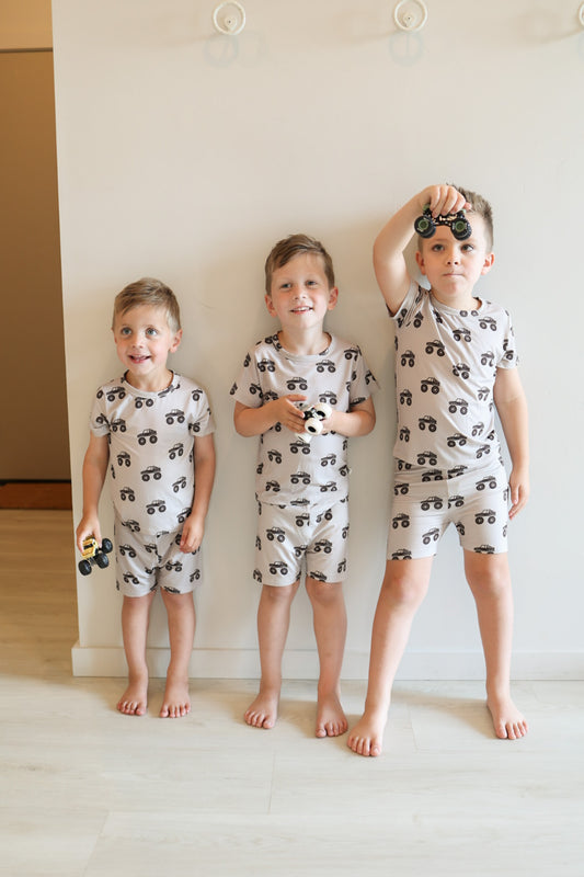 Three barefoot boys stand by a light wall, wearing forever french baby Bamboo Short Set | Monster Truck pajamas. Two hold toy cars, while the tallest lifts his car overhead, enjoying their comfy, breathable sleepwear.