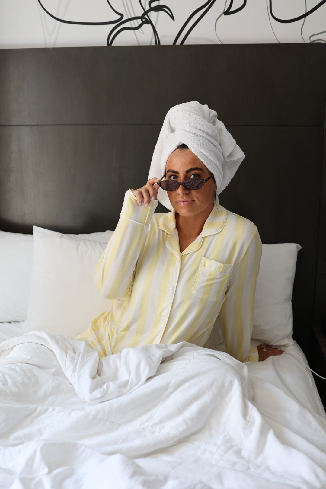 A person in Long Sleeve Women's Bamboo Pajama | Butter by forever french baby sits on a bed with a white comforter and pillows, wearing a towel wrapped around their head and lifting sunglasses to their face in a modern bedroom.