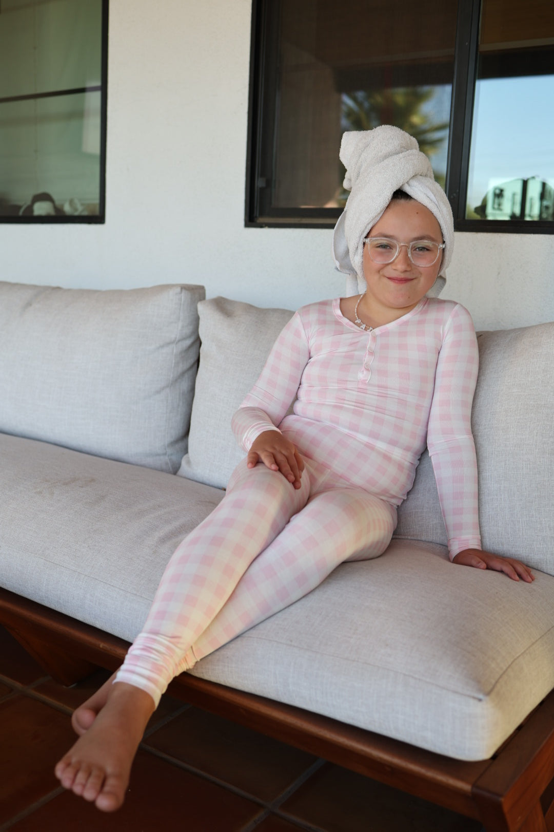 A girl in forever french baby’s Bamboo Two Piece Pajamas | Pink Sugar, with a towel on her head, sits on a couch enjoying cozy, hypo-allergenic comfort.