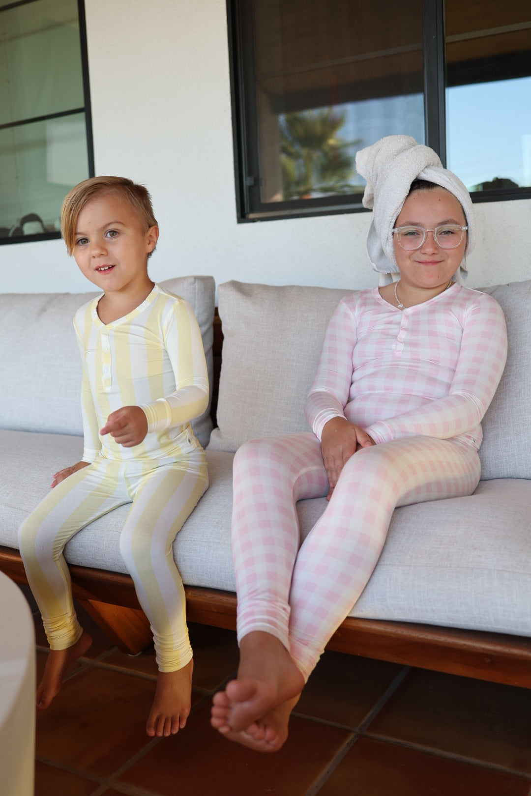 Two children lounge outside in forever french baby’s Bamboo Two Piece Pajamas. One wears yellow-and-white stripes; the other, Pink Sugar pajamas, with a towel-wrapped head and glasses. Both look relaxed and comfy in their breathable kids sleepwear.