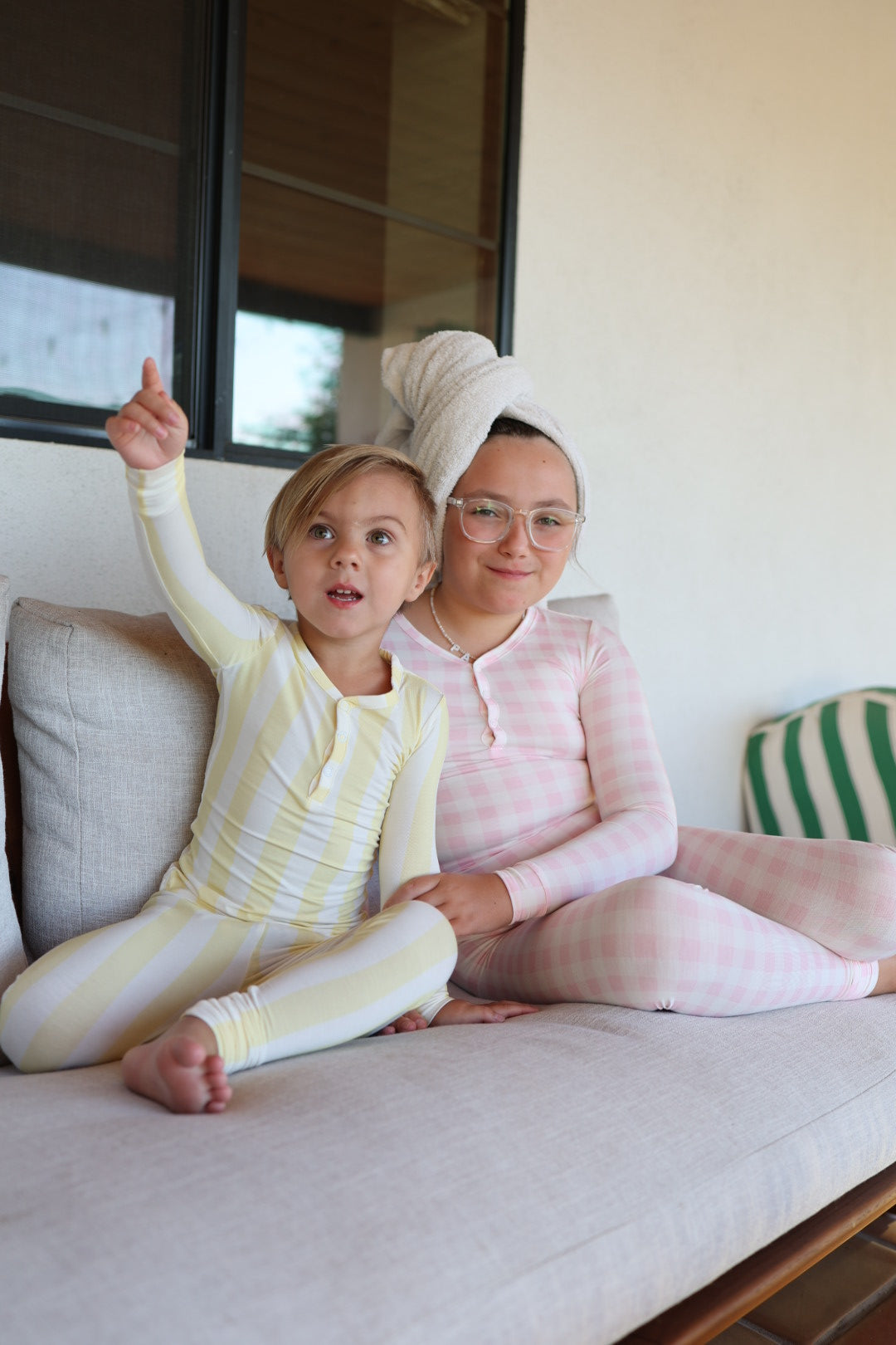 Two children sit on a cushioned bench. One, in yellow-and-white sleepwear, points and smiles, while the other grins at the camera in forever french baby’s Bamboo Two Piece Pajamas | Pink Sugar, glasses, and a towel turban.