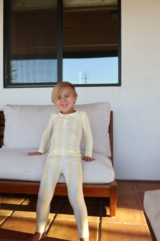A young child with blonde hair smiles while sitting on a cushioned outdoor bench, wearing forever french baby’s Bamboo Two Piece Pajamas in Butter—yellow and white stripes—against a white wall and window backdrop.