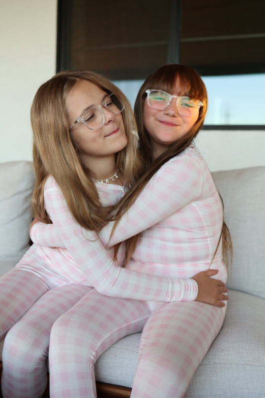 Two young girls wearing forever french baby Bamboo Two Piece Pajamas in Pink Sugar and glasses sit on a couch, hugging each other and smiling happily.
