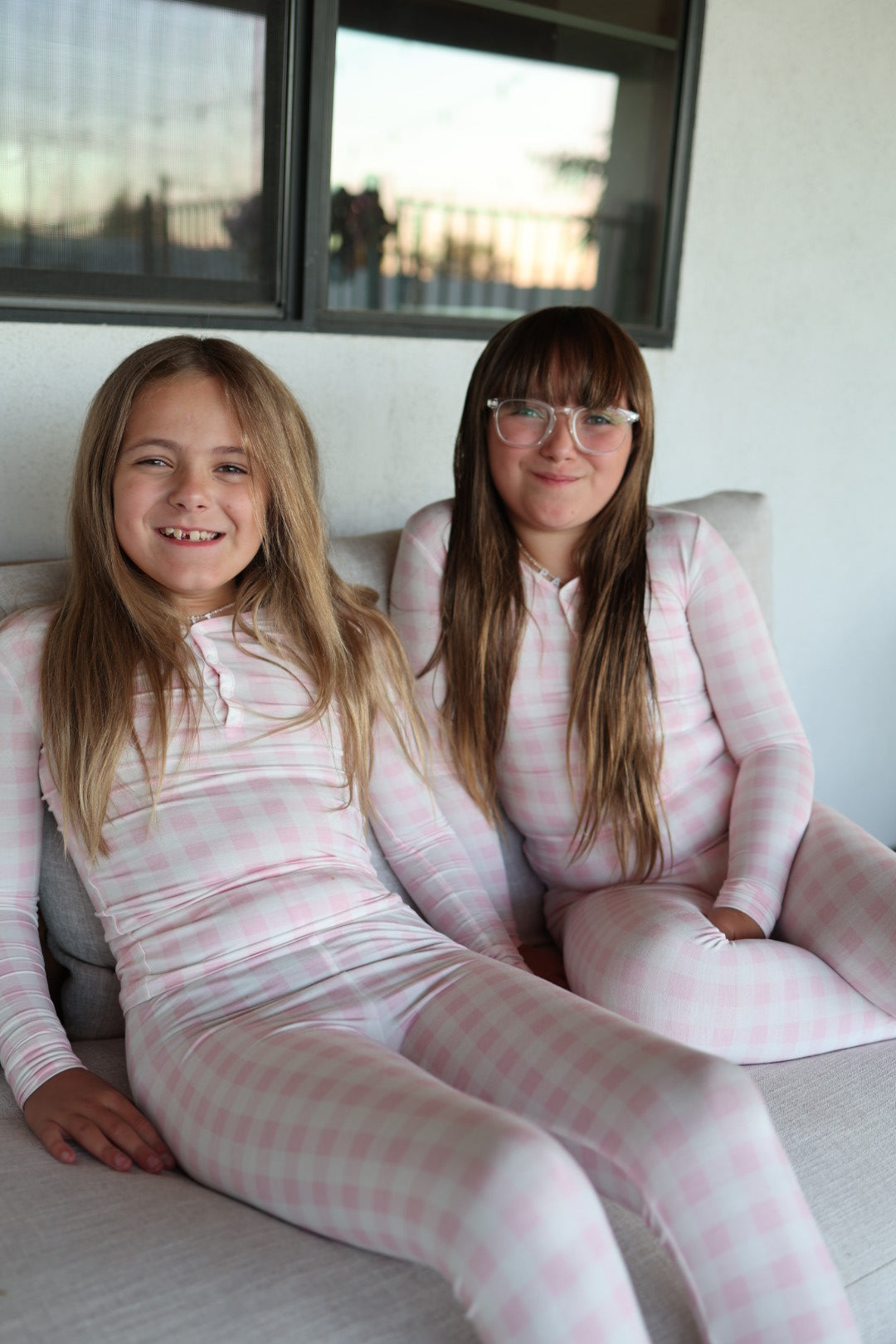 Two young girls with long hair wear matching forever french baby Bamboo Two Piece Pajamas in Pink Sugar as they sit together on a light sofa, smiling at the camera. A window is visible behind them.