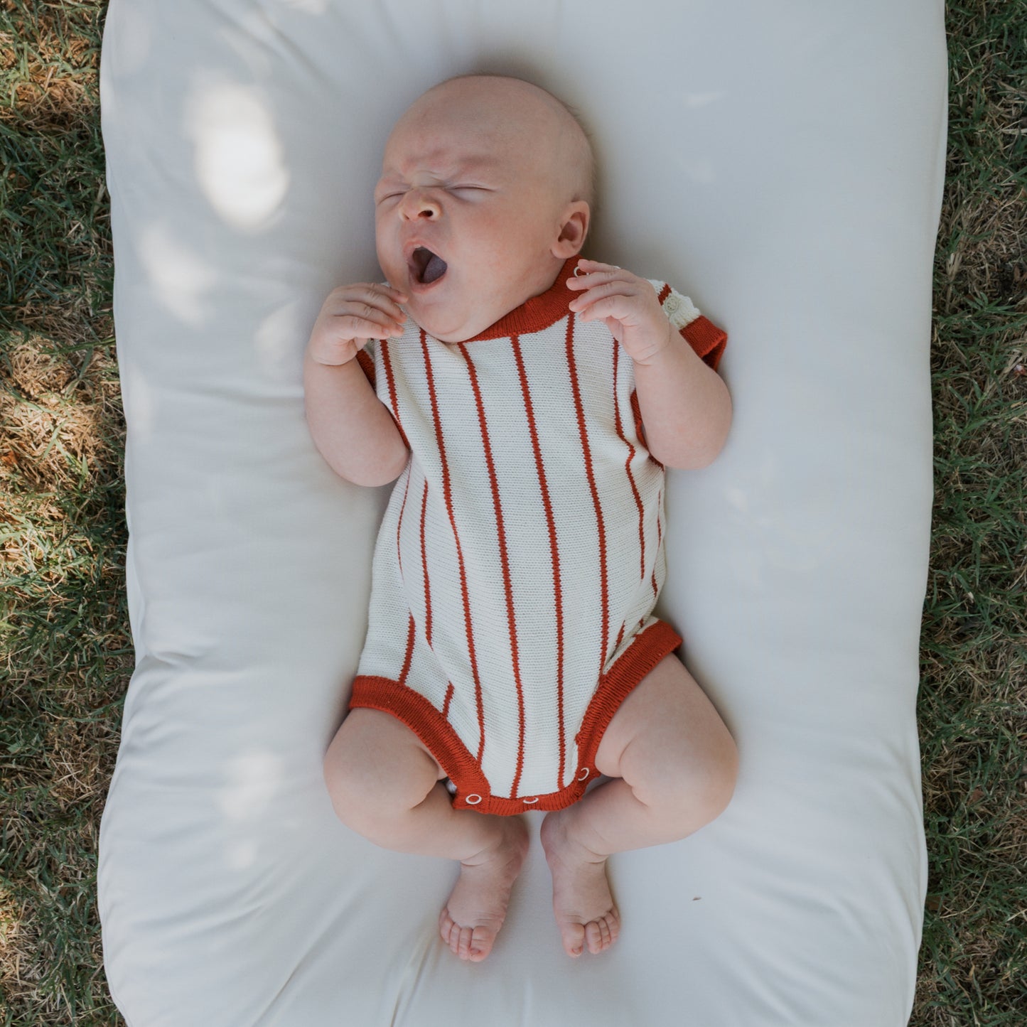 A baby in the forever french baby Knit Romper | Picnic Pinstripe, made from 100% cotton, lies on a white cushion outdoors with hands near the face, yawning peacefully as green grass surrounds the cozy scene.