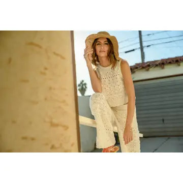 A woman wearing MIOU MUSE's Crochet Pattern Pants and a matching cotton crochet top sits on an outdoor ledge, accessorized with a wide-brimmed hat. The backdrop displays a building against a clear sky, suggesting a sunny day. Her delicate attire is best maintained with hand wash care.