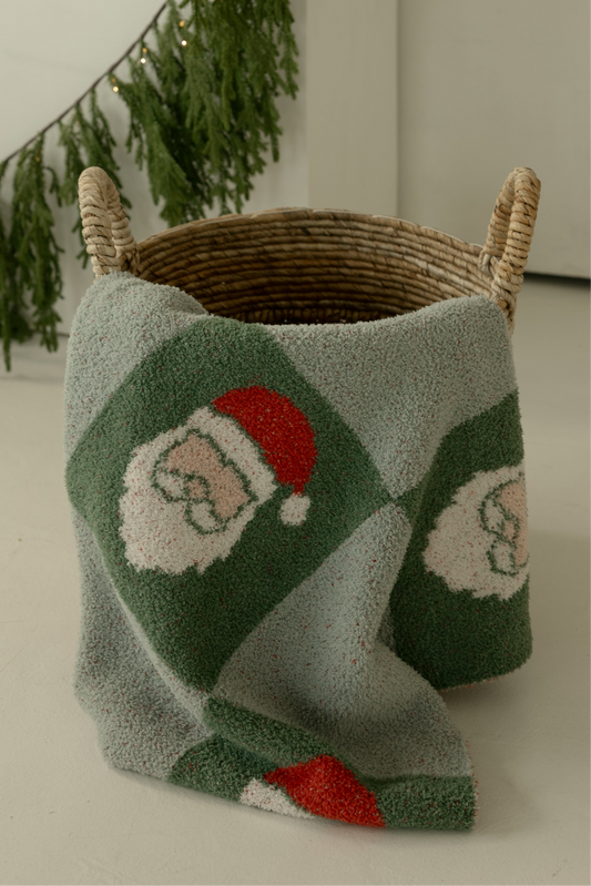 A woven basket holds the forever french baby Plush Blanket | Santa Checkerboard in green and white, featuring Santa Claus faces and draped over the side. Pine garland in the background adds a festive touch.