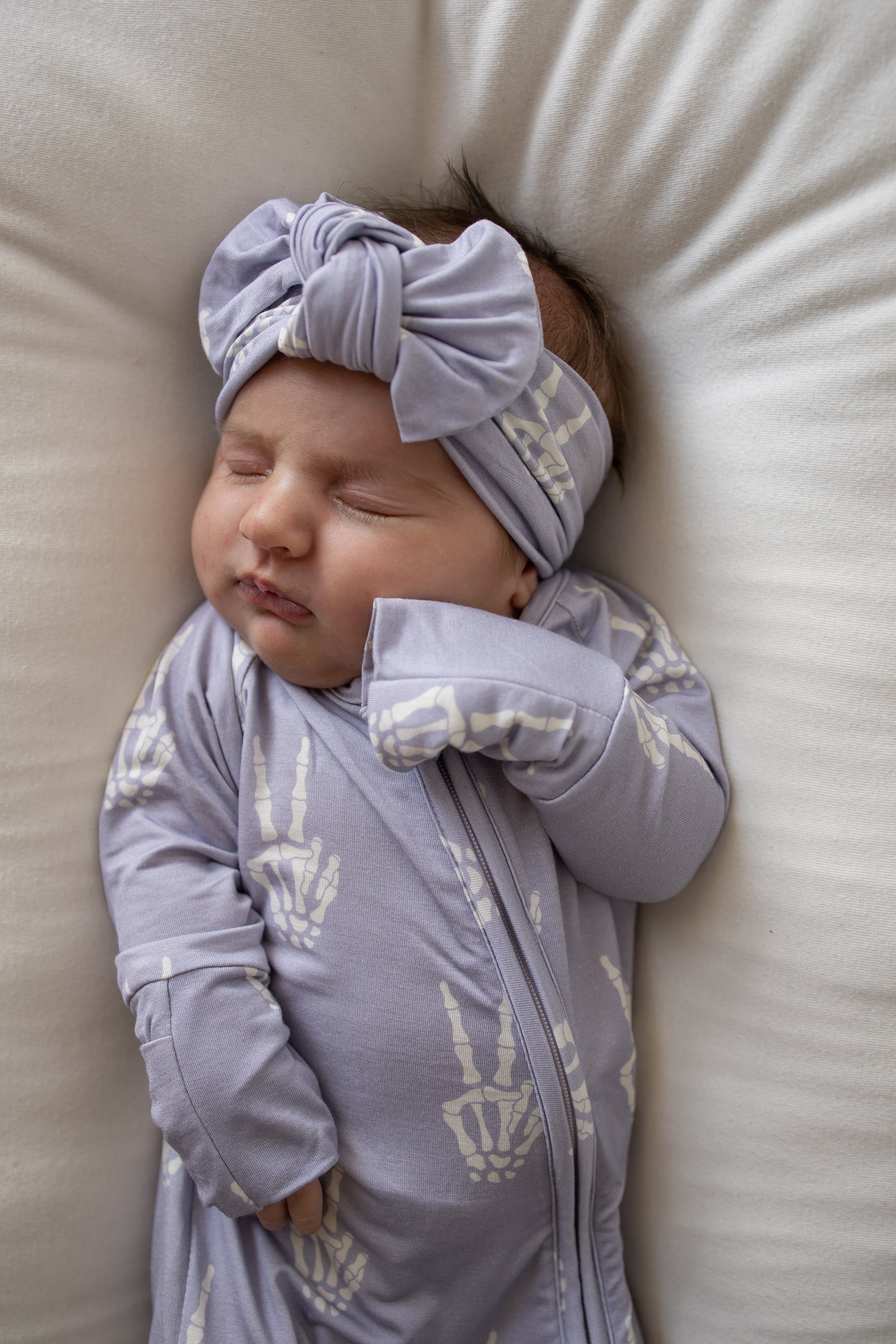 A sleeping baby wearing the Forever French Baby Bamboo Head Wrap | Enchanted Skelly and a matching onesie with white skeleton hand prints rests on a white cushion, highlighting the adorable style of forever french baby.