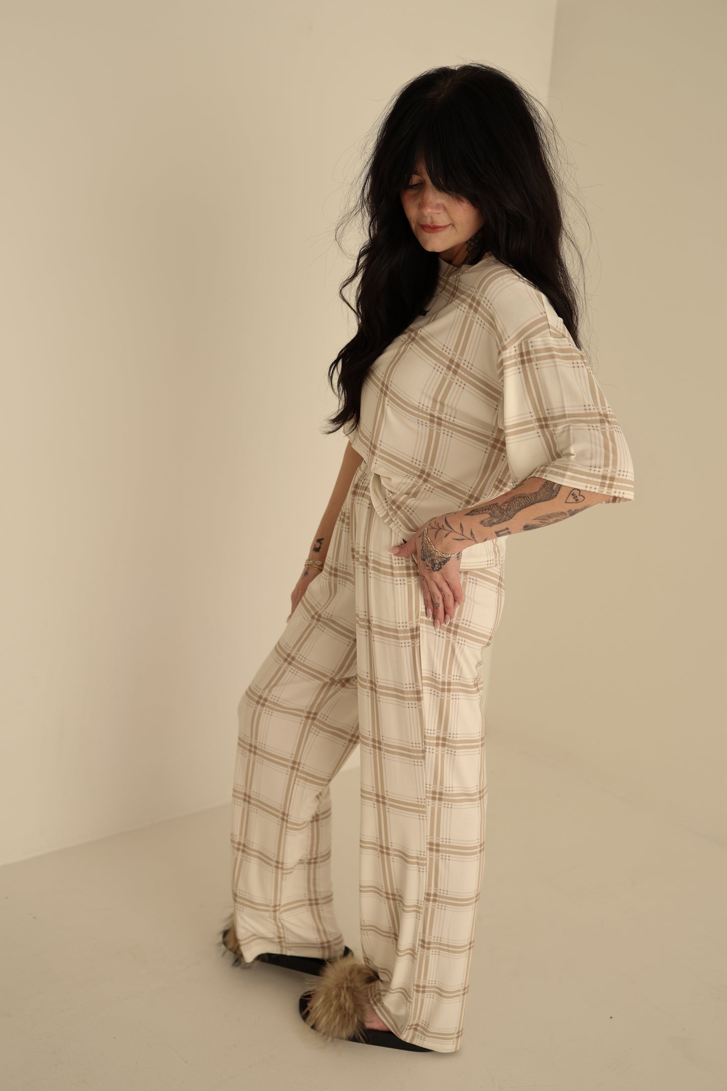 A woman with long dark hair and tattoos models the forever french baby Women's Boxy Tee Lounge Set in Ellis Plaid, featuring wide-leg pants. She stands in a minimalist room, pairing the cozy cream set with furry slippers for a stylish look.