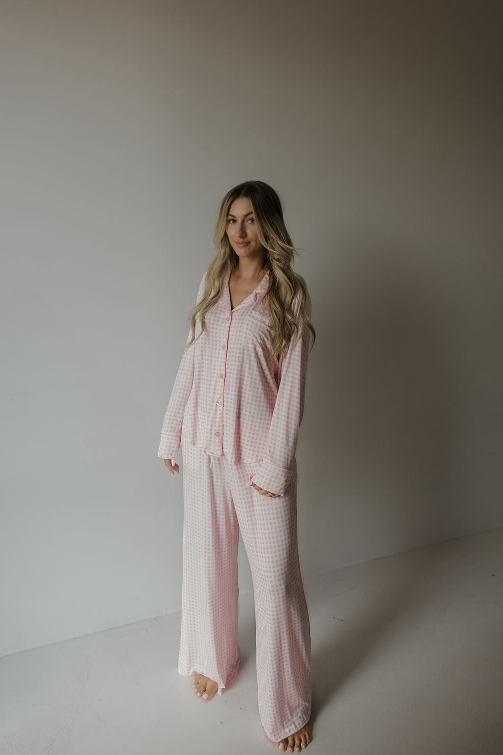 A woman stands in a minimal room, barefoot, wearing the forever french baby Women’s Bamboo Button Up Lounge Set | Giselle—a loose-fitting, light pink and white checkered pajama set with wide-leg lounge pants and long sleeves. Her long, wavy blonde hair cascades over her shoulders.