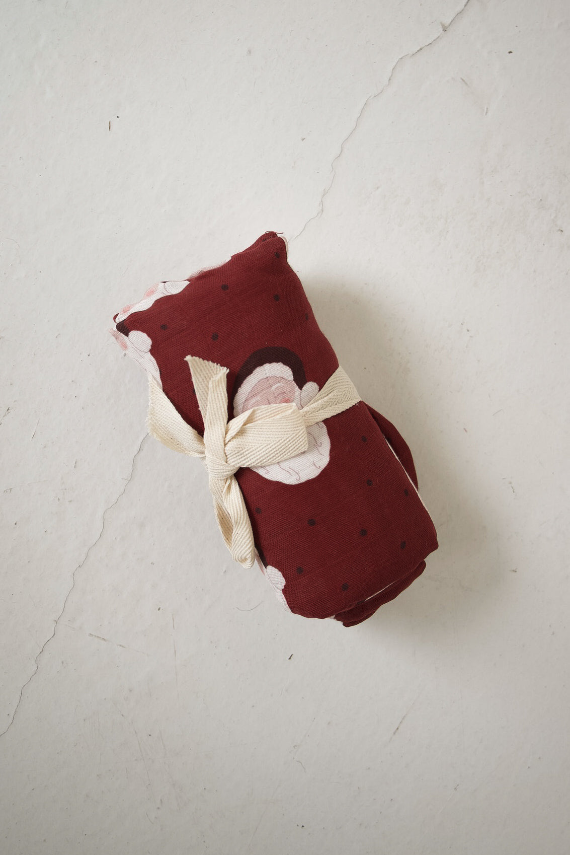 A dark red forever french baby Muslin Swaddle | Vintage Santa featuring a vintage Santa and polka dot pattern, rolled and tied with a cream ribbon.